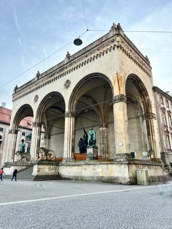 A large stone pavilion with delicate trim holds three statues, two copper and one bronze, two lions stand guard alongside the steps, two pedestrians walk across the square under a blue sky with wispy cloud.