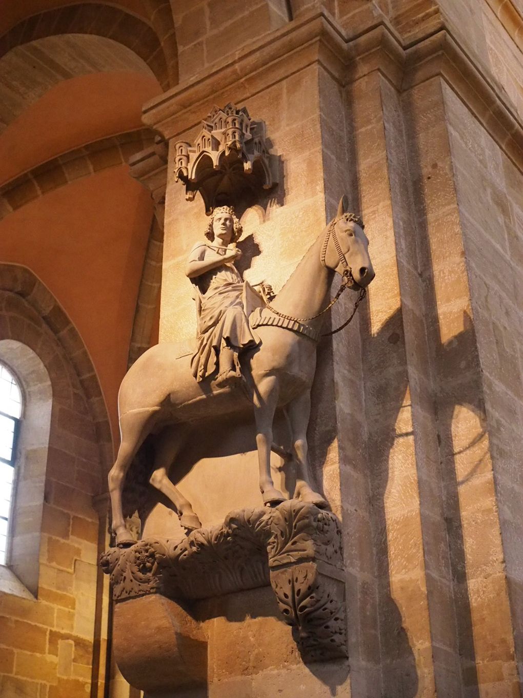 A large tan statue of a mounted knight on a horse is affixed to the side of a column in a cathedral, he is wearing a crown and gazes serenely over the viewer's head, the horse is shown lifesized.
