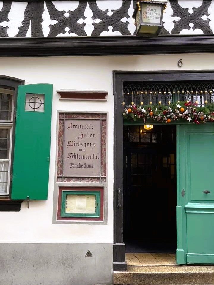 The entrance to an old inn with a green door, green shutters and a Christmas decorations is shown. The dark beams and timber fretwork are shown, a sign on the walls states that its the brewey heller, inn called Schlenkerla.