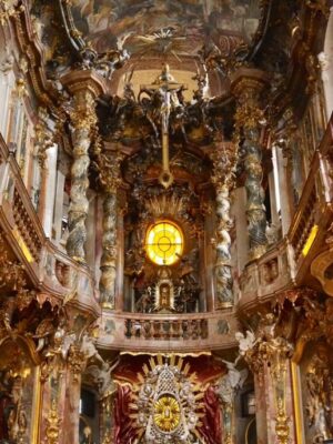 A heavily decorated chapel interior practically drips with gold, embellished with gilt carvings and statues, the eye is drawn to a golden window suspended above the altar, a highlight of the Munich 3 day itinerary