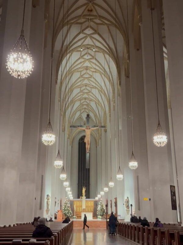 A forest of thick white columns, topped by timber framing, lead the eye to a large crucifix and high altar, lights hang fromthe ceiling and a lone man walks at the end of the aisle. Parishioners dressed in black dot the pews.
