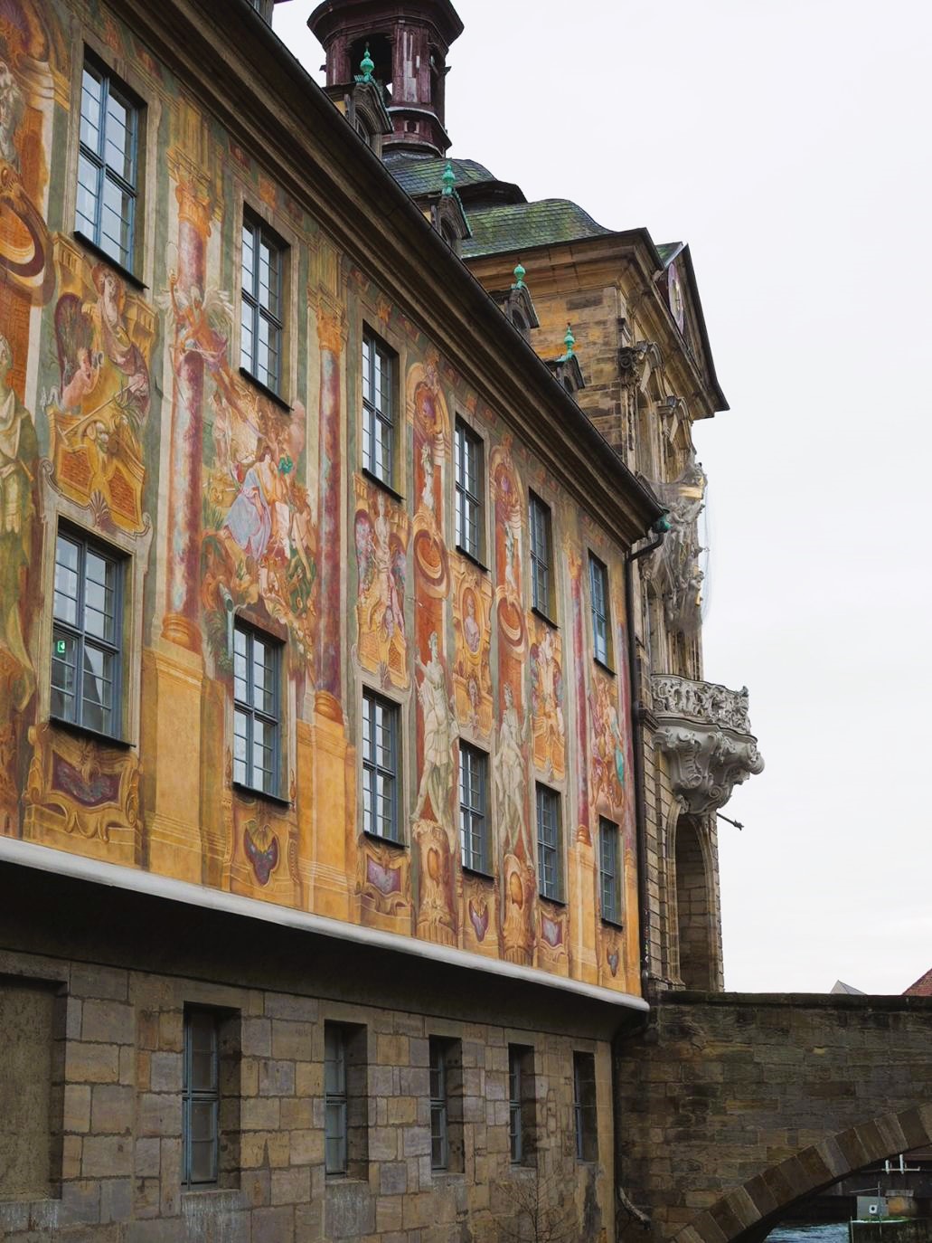 The side of a Gothic building is shown covered in vibrant frescoes displaying robed figures amidst decorated columns, a footbridge is connected to the building