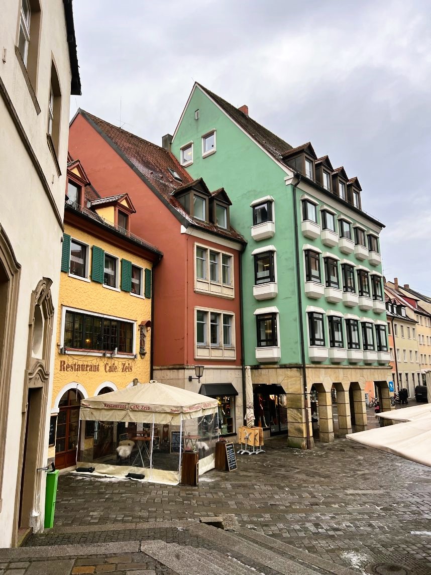A small cobblestoned square is shown ringed by quaint high roofed houses, decorated in shades of yellow, red and green, windows peep out from the roofs.