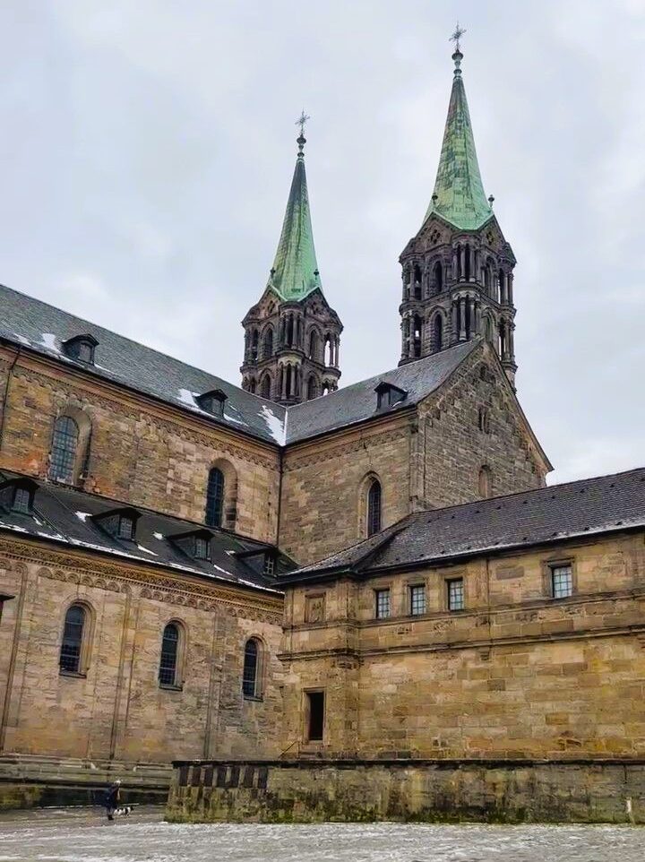 Sandstone blocks for the walls of a cathedral and a palace attached to it, the dark roofs are dusted with snow and twin copper spires rise above the scene against a cloudy sky.