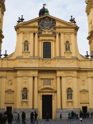A large Baroque church stands on a city square, painted in a vibrant yellow hue, statues adorn the facade and a grey Bavarian coat of arms crests the building
