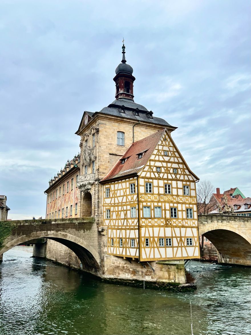 A gothic town hall, made of sandstone, is situated in the middle of the Regnitz river in Bamberg, adjoined on either side by bridges. A yellow timbered house is attached at the end closest to the viewer.