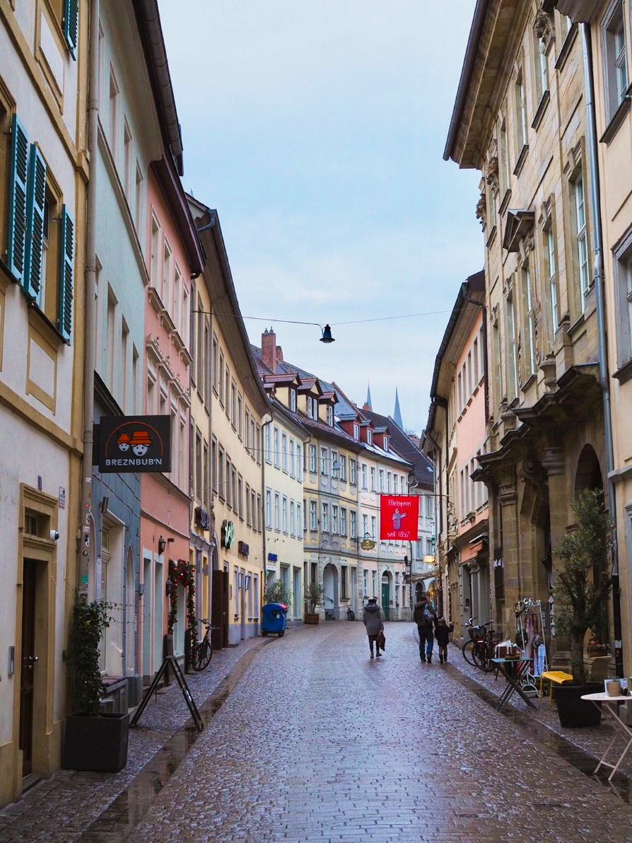 A cobblestoned street lined with pastel coloured townhouses curves gently away from the viewer, sparse visitors peruse the shops at street level.