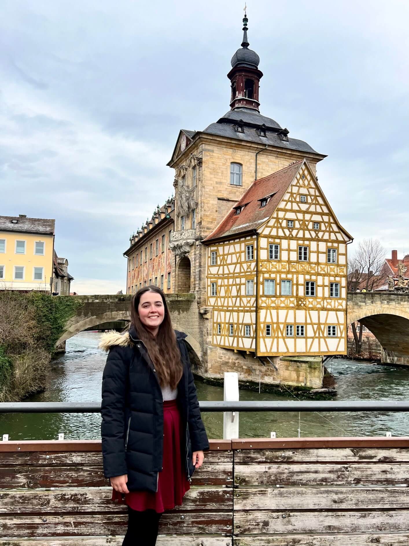 A lady in a puffer jacket and red skirt with her brown hair out stands in front of a Gothic town hall located in the middle of a river, bridges join it on either side.