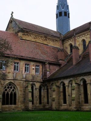 A green grassed courtyard is surrounded by sandstone buildings, most with red tiled roofs and a dark church steeple rising from the corner of the buildings.