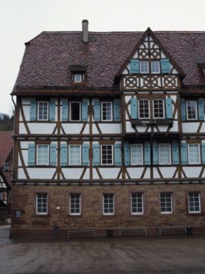 The large half timbered building of the Bursarium stands tall against a cloudy sky, its blue shutters and dark timber details contrast against its sandstone foundation