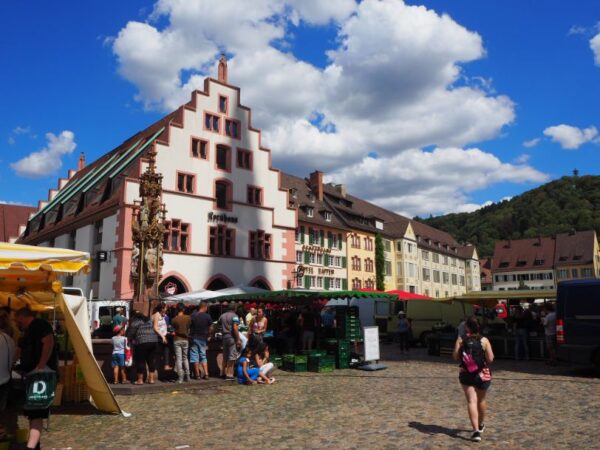 The sun shines brightly over a bustling market square, surrounded by impressive townhouses, one with a stepped roof. Market stalls crowd the cobblestones while visitors browse the wares. The sky above is studded with white, fluffy clouds.