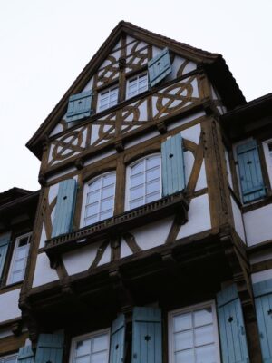 A large bay window stands out from the facade of a half timbered house, adorned with light blue shutters and charming wood framing.