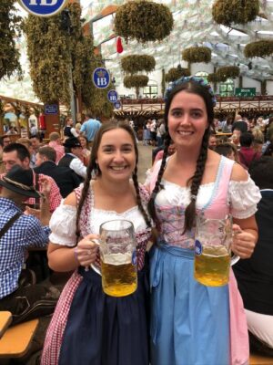 Two ladies dressed in traditional Bavarian dirndls hold steins of beer, their dresses are red checked and blue and pink, with blue aprons. Their hair is in plaits and they stand in a large Oktoberfest tent