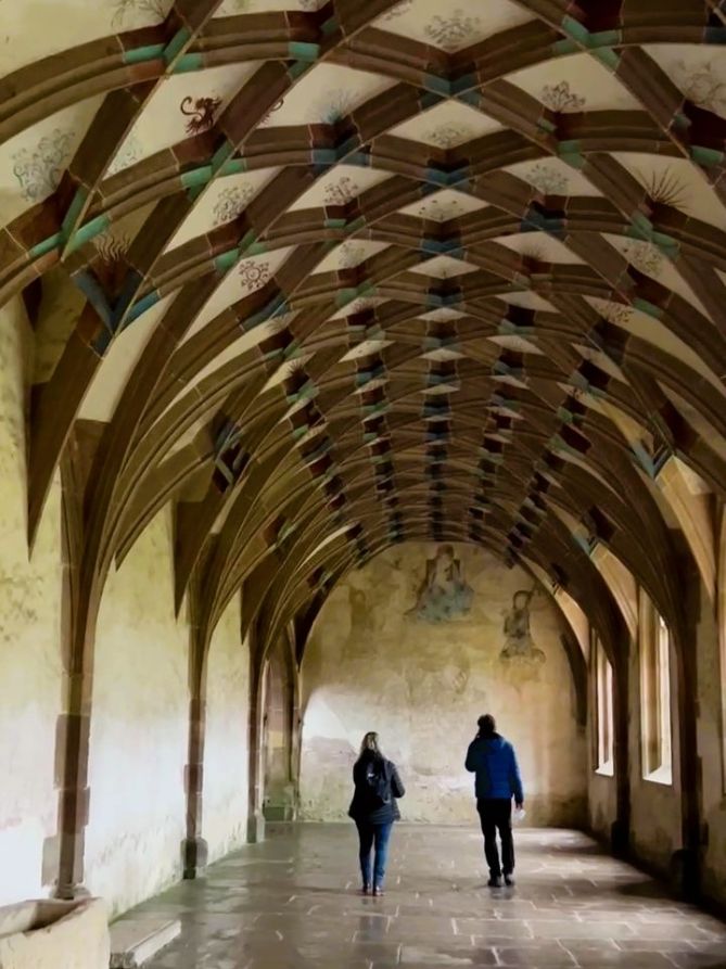 Two people in puffer jackets stroll beneath an ornate ceiling of chestnut arches, accented with blue paint against white plasters. Including red and blue illustration on the roof, and murals at the end of the hall. Light streams in from windows at the side.