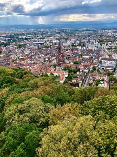 A large city centre, complete with a red roofed Old Town and cathedral spire, stands in a valley in the Black Forest. Rain clouds gather in the distance while many green trees cluster in the foreground.