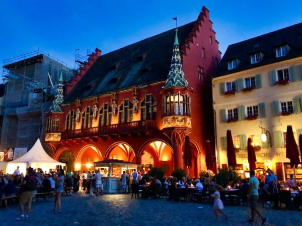 A large, scarlet coloured merchant's hall, topped with two elegant turrets at each end, stands on a city square. Its arches are lit up with lights as crowds of people sit at large tables and enjoy the evening summer air.