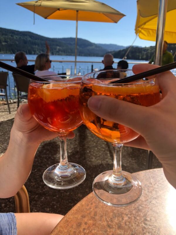 Two hands are holding up red Aperol Spritzes in wine glasses topped with straws and slices of orange in front of a patio filled with umbrellas and people watching a lake in the background.