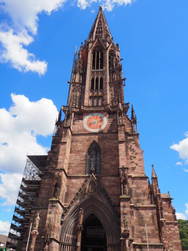 A lacework inspired red sandstone tower reaches up to a blue sky, studded with clouds. A large clock with a red face stands high on the facade above intricate portal doors. Some scaffolding is visible low on the left hand side.