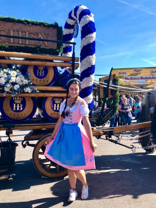A lady is dressed in a pink and blue traditional Bavarian dirndl, with her dark hair in plaits. She stands in front of a wooden wagon filled with barrels of beer and a wreath covered in white and blue streamers.