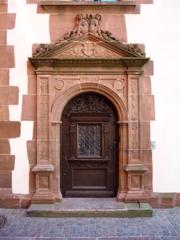 A large pink sandstone door frame, heavily decorated in intricate carvings, including two naked angels atop the frame, surrounds an equally ornate dark wood door, inset with glass. The street is lined with cobblestones.