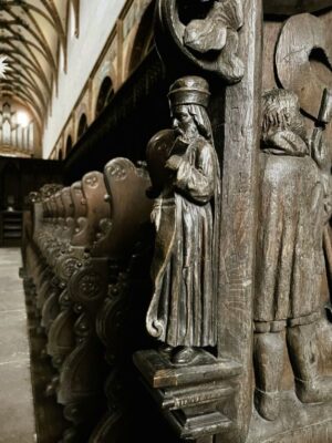 A little man is hand carved into the end of dark wood choir stalls, including flowing robes and an elaborate hat. Even his little boots. The rest of the church stretches away in the distance including delicate arches of stone for the ceiling
