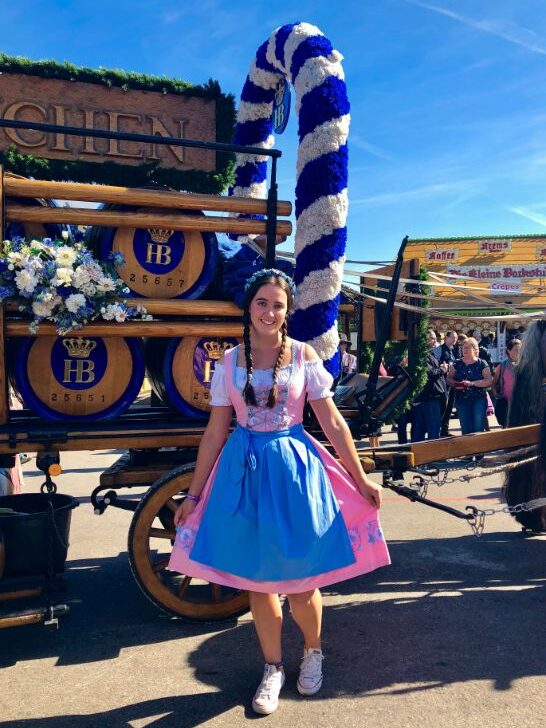 A lady is dressed in a pink and blue traditional Bavarian dirndl, with her dark hair in plaits. She stands in front of a wooden wagon filled with barrels of beer and a wreath covered in white and blue streamers. She looks at the camera smiling
