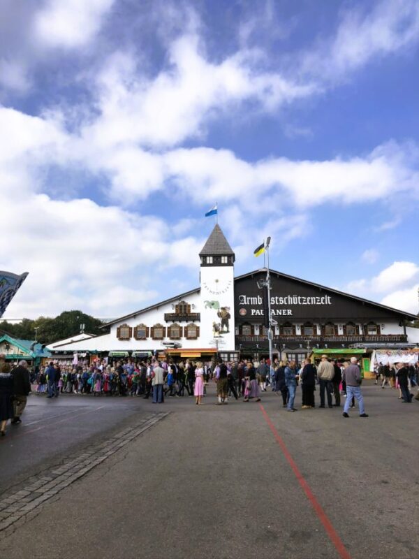 A large buildng with a clock tower is shown beneath a blue sky dotted with clouds, a group of people stand before it, the text on the building reads Armbrustschützenzelt and Paulaner for the beer company