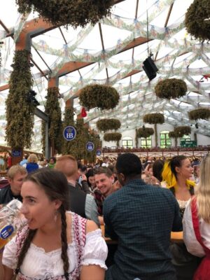 Young people line the tables of the HB tent at Oktoberfest in Munich, a lady in a checkered dirndl drinks out of a glass stein, HB signs and hop garlands hang from the roof. Find views like these in the ultimate guide of where to stay in Munich for Oktoberfest.
