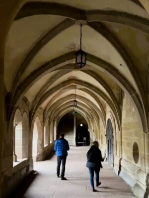 Two peope dressed in blue and black puffer jackets walk the sandstone hallway of the Paradise in Maulbronn, elegant arches frame the space and daylight streams in through gently curved portals