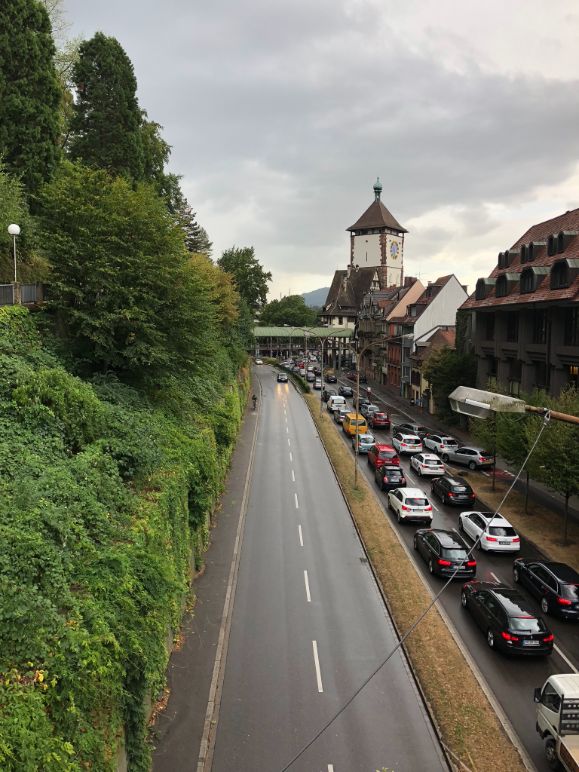 Four lanes of grey tarmac, the right side filled with cars sit between luscious green hills and the elegant townhouses that form the edge of Freiburg's old town. The Schwabentor tower is visible in the distance under a cloudy sky
