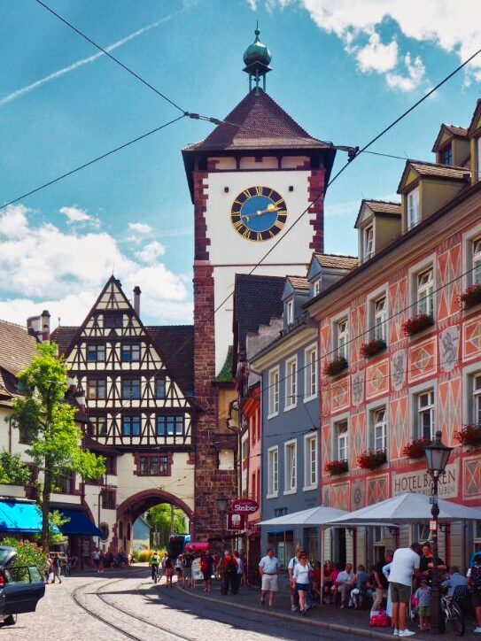 A large red sandstone and white plaster tower stands tall over the old town of Freiburg. A blue clock is high on the facade and its surrounded by charming multi-coloured town houses, while tram tracks curl along the city streets. The sky above is blue with dappled cloud and visitors stick to the shade offered by restaurant umbrellas