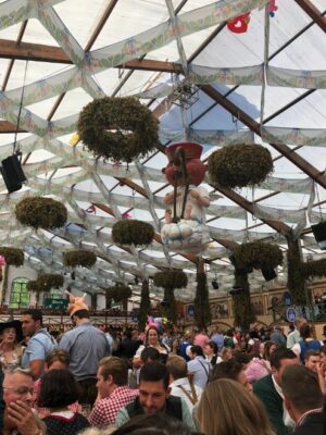 A crowd is shown seated at tables while above, garlands of green hops and a large, red faced angel sits on a cloud with a harp.