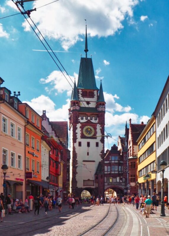 A large Gothic tower, topped with multiple copper spires and a blue green roof, stands at the end of a gently curving city street, surrounded by multi coloured townhouses and bathed in brilliant sunshine