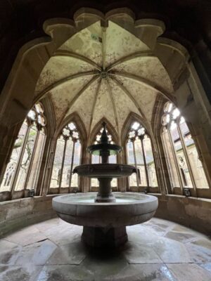 A rounded fountain is shown within its ornate fountain house, complete with archwork and a flagstone floor, the fountain has three distinct tiers and a bronze topper, the windows are ornate with stonework and illustrations cover the ceiling panels at Maulbronn Monastery