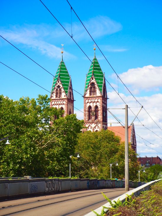 Two large church spires, decorated with emerald green roofs and white and brown contrasting stonework stand against a blue sky studded with white clouds. Tram lines run through the scene alongside green trees.