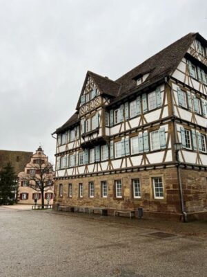 A view over a large, handsome half timbered building with a light stonework foundation and stucco and timber work upper stories, as well as a bay window. The shutter are robin egg blue and we can see the pink Maulbronn Rathaus in the distance, above the sky is cloudy