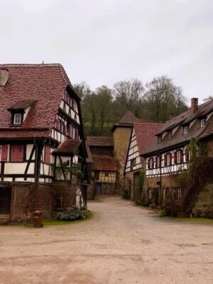 Multiple half timbered buildings with roofs and shutters in shade of red stand over a courtyard of light coloured stones, damp from the rain beneath a cloudy sky. Bare branched trees reach up the nearby hillside.