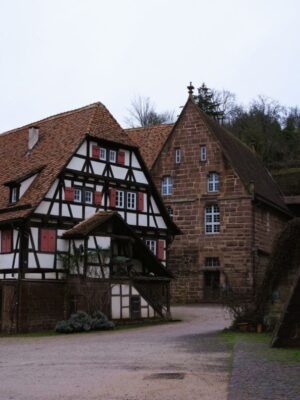 A large four storied house with red shuttered windows and a tiled roof stands before a stonework building which used to house the monastery's flourmill. The ground is wet from recent rain and the sky above is cloudy over bare branched trees.