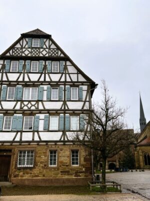 A large, five storied house with half-timbered accents and blue shutters stands under a wintry sky, the monastery church spire can be seen in the background