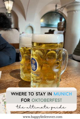 Two golden foam topped steins of beer stand on a wooden table, both have the Hofbrauhaus logo on them. The text reads where to stay in Munich for Oktoberfest the ultimate guide