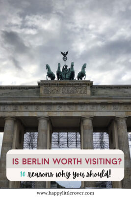 The famous Brandenburg Gate stands against a dramatic cloudy sky, the four blue-green copper horses lead a chariot atop the thick stone crossbeam of the gate. Scaffolding can be seen behind the thick columns. The text reads is berlin worth visiting, 10 reasons why you should.