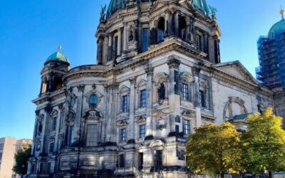 The copper domed Berlin Cathedral rises majestically above the green River Spree, sunlight dapples the green trees next to the cathedral and the whole scene is topped by a bright blue sky
