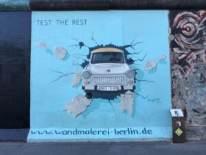 A light blue mural is painted onto the remnants of the Berlin Wall, accented with a light coloured car depicted as breaking through the wall with its numberplate reading Nov.9.89, a small informational sign stands to the right of the mural and the heading in the top left reads Test the Rest.