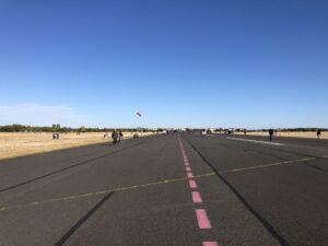 A wide tarmac runway bisected by yellow and pink lines stretches off into the distance, visitors stroll along the yellowed grass and dark asphalt, a kite surfer's kite can be seen in the distance against a blue sky.