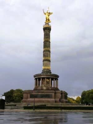 A large, dramatic column studded with golden accents and topped with a golden statue of Victory stands against a stormy sky, a ring of columns surrounds the base of the monument.