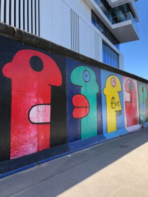 Large, vibrantly coloured humanoid figures feature on a mural painted onto the former Berlin wall which stretches away from the viewer, an apartment block stands above it under a blue sky.