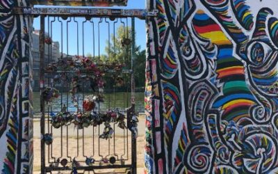 Sunlight streams through a wire gateway adorned with padlocks, the wall surrounding the gateway is covered in vibrant, rainbow murals in dramatic swirls.