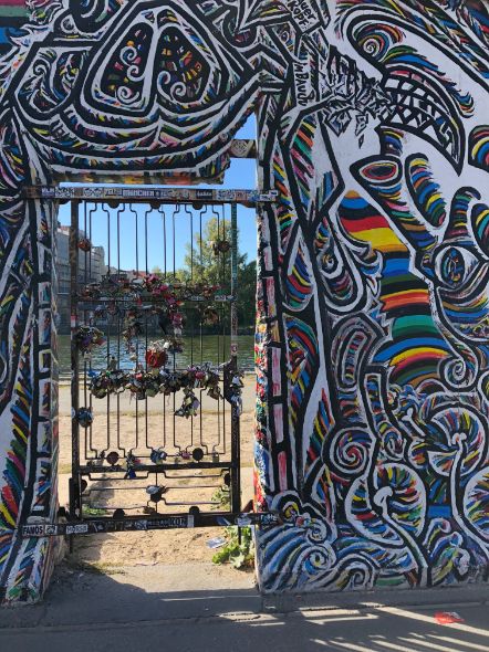 Sunlight streams through a wire gateway adorned with padlocks, the wall surrounding the gateway is covered in vibrant, rainbow murals in dramatic swirls.