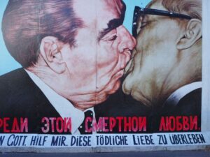 Two suited men are painted in great detail kissing on a light blue background painted onto the remnants of the Berlin Wall in Berlin with red Cyrillic script and black German writing below that.