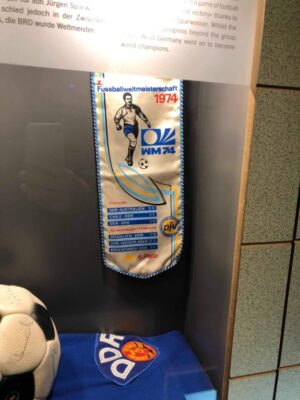 A small blue, white and yellow pennant is affixed to a grey wall and displays a soccer player kicking a ball and various fixtures results from the former DDR soccer team, beneath a dark blue soccer jersey and signed soccer ball are resting within the museum exhibit.